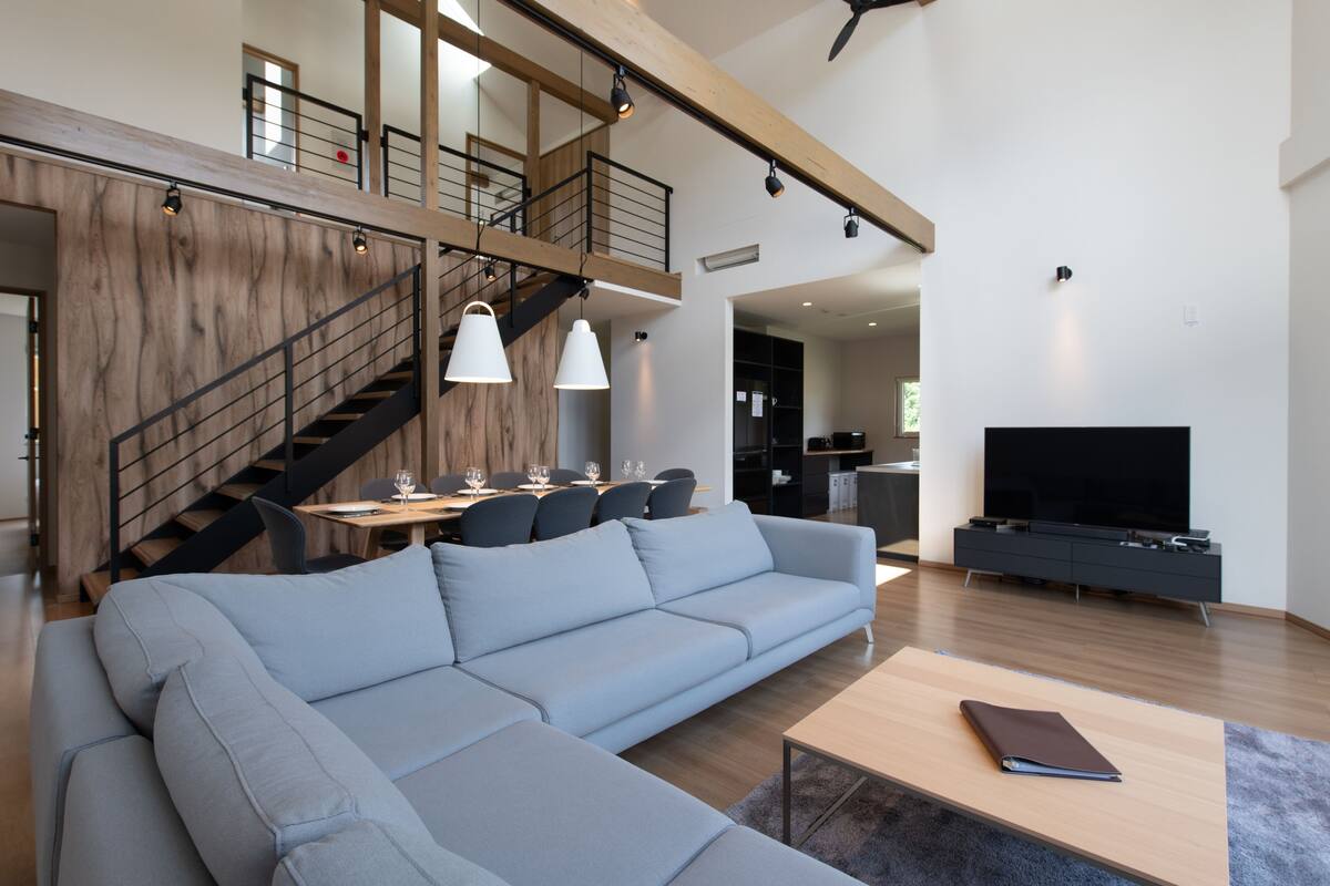 A spacious living area with a large grey sectional sofa, a wooden coffee table, and a flat-screen TV on a black stand. A staircase with wooden railings leads to an upper level.