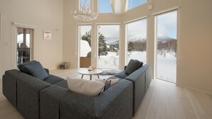 A modern living room with a grey sectional sofa, a small round table, and a chandelier. Large windows offer a view of a snowy landscape. - The Orchards Niseko (Kutchan)