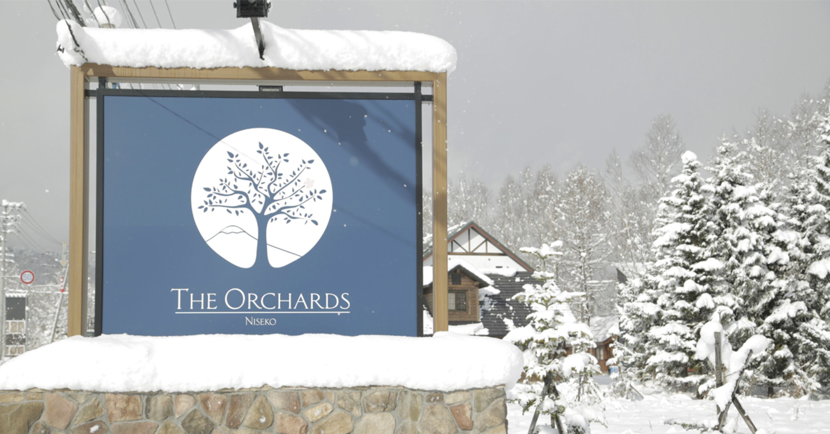 Skilt for "The Orchards" med et trælogo, mod en sneklædt natur med huse i baggrunden.