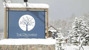 Skilt for "The Orchards" med et trælogo, mod en sneklædt natur med huse i baggrunden.