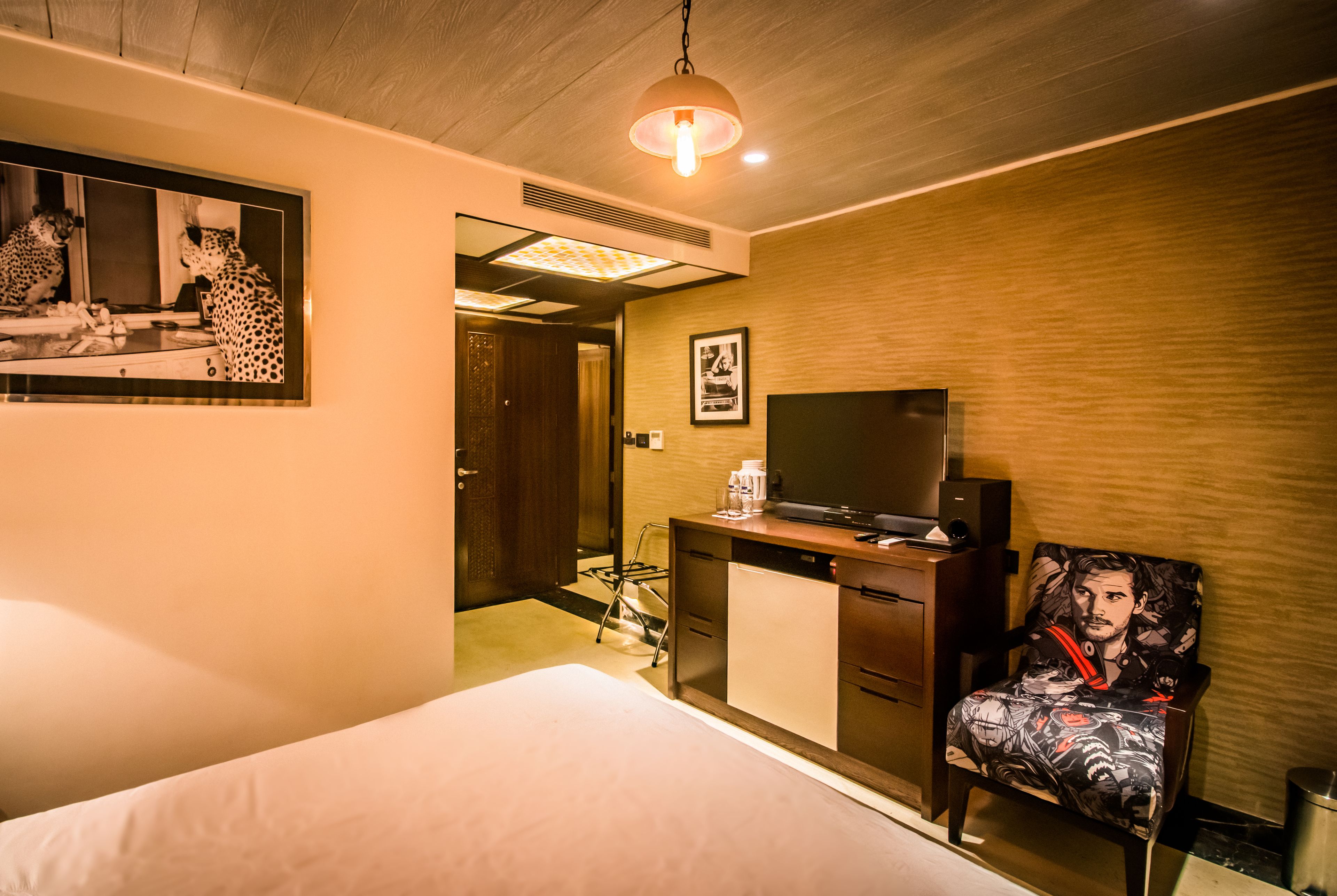 A hotel room with a bed, a desk with a television, a chair with a graphic design, and a mirror on the wall.