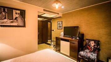 A hotel room with a bed, a desk with a television, a chair with a graphic design, and a mirror on the wall.