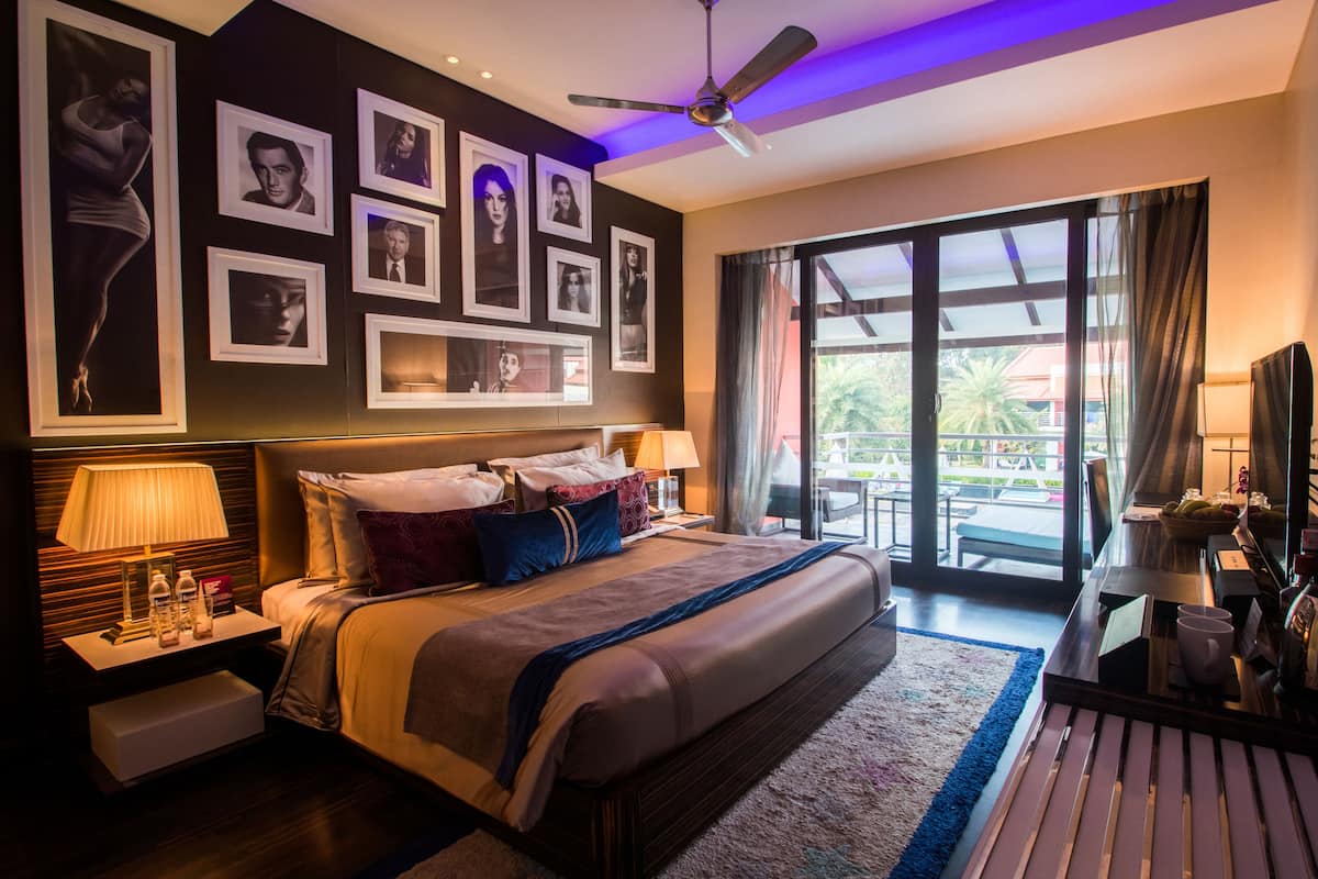 Habitación de hotel con una cama grande, ventilador de techo, mesita de noche con lámpara, escritorio con computadora y balcón con vistas.