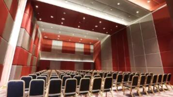 Un salón de conferencias con filas de sillas, piso a cuadros y un escenario con un fondo rojo y blanco.