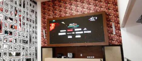 Una habitación con un televisor de pantalla plana grande montado en una pared con papel tapiz floral, suelo de madera y una pared llena de fotografías enmarcadas.