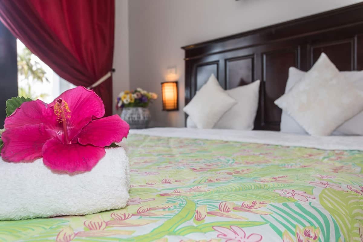 Een hotelkamer met een bed, een bloemenlaken, een witte handdoek met een hibiscusbloem en een nachtkastje met een bloevas.