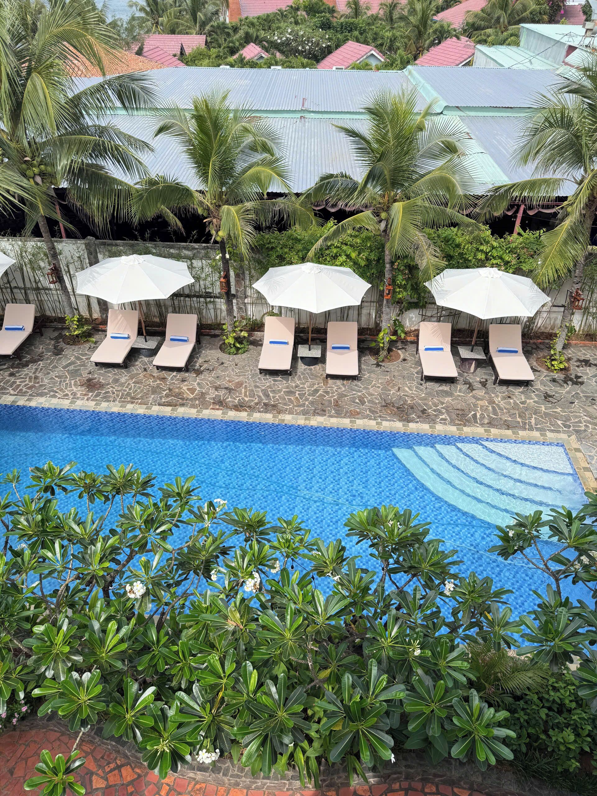 A pool area with lounge chairs and umbrellas, surrounded by lush greenery and a view of buildings with red roofs.