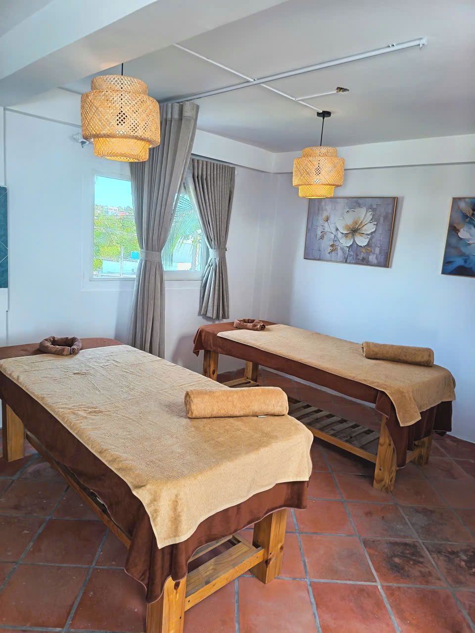 A room with two wooden massage tables, a window with curtains, and two wall-mounted lamps.