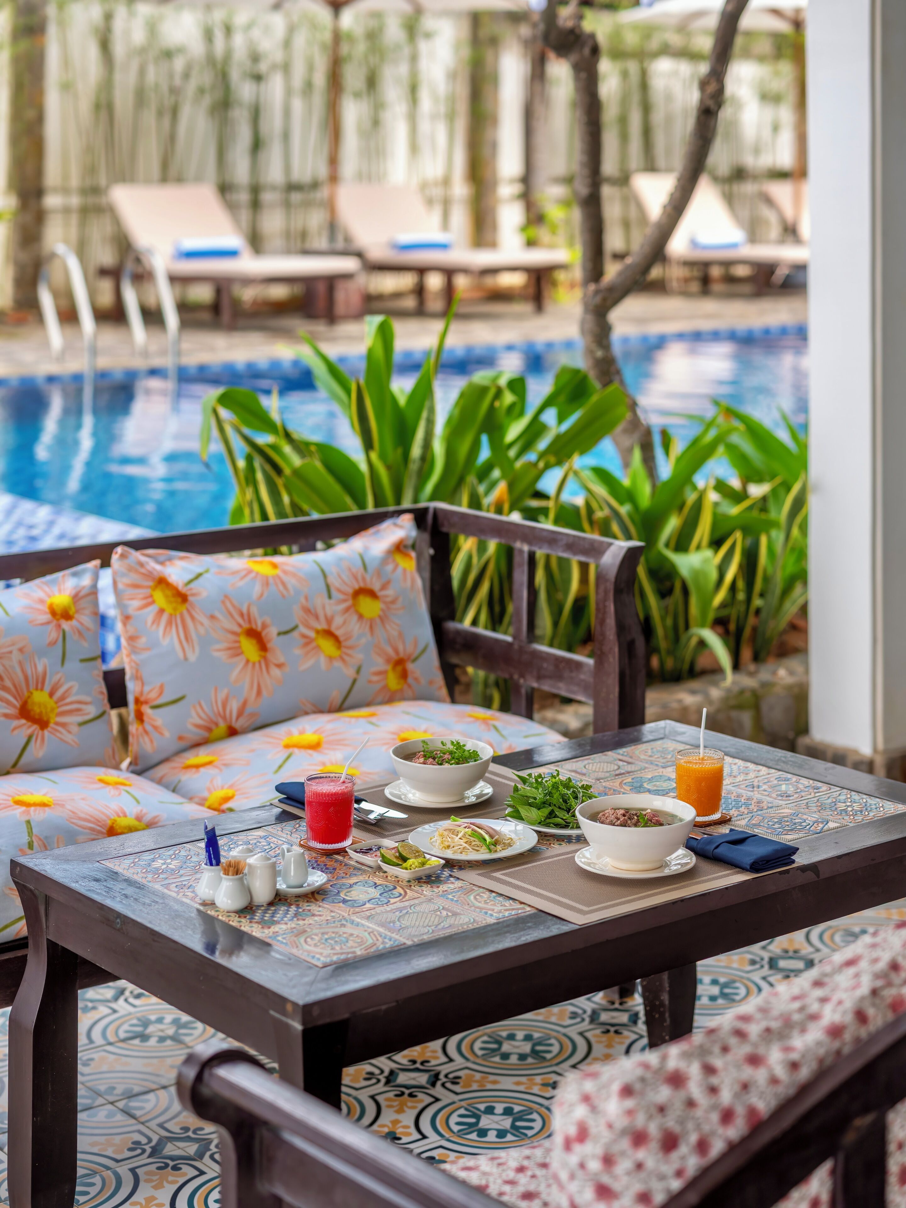 A dining table with a floral cushion, a bowl of soup, a plate of noodles, a glass of juice, and a napkin.