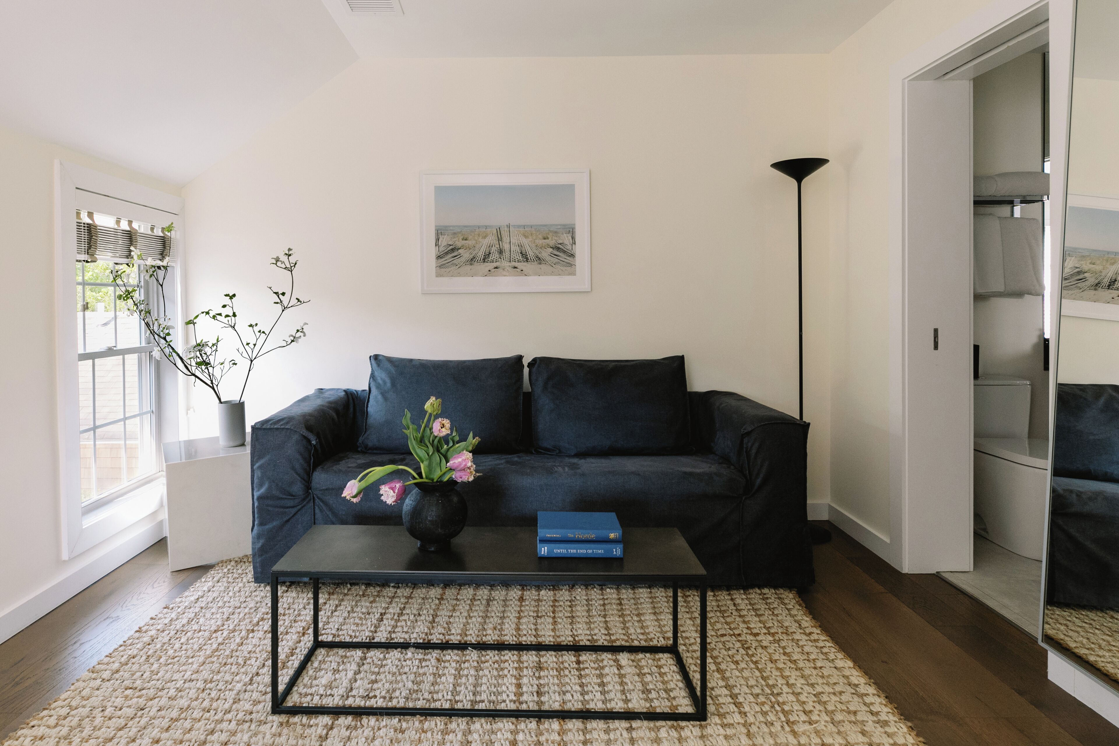 A living room with a dark blue sofa, a black coffee table with a vase of flowers, a framed picture on the wall, and a tall floor lamp.