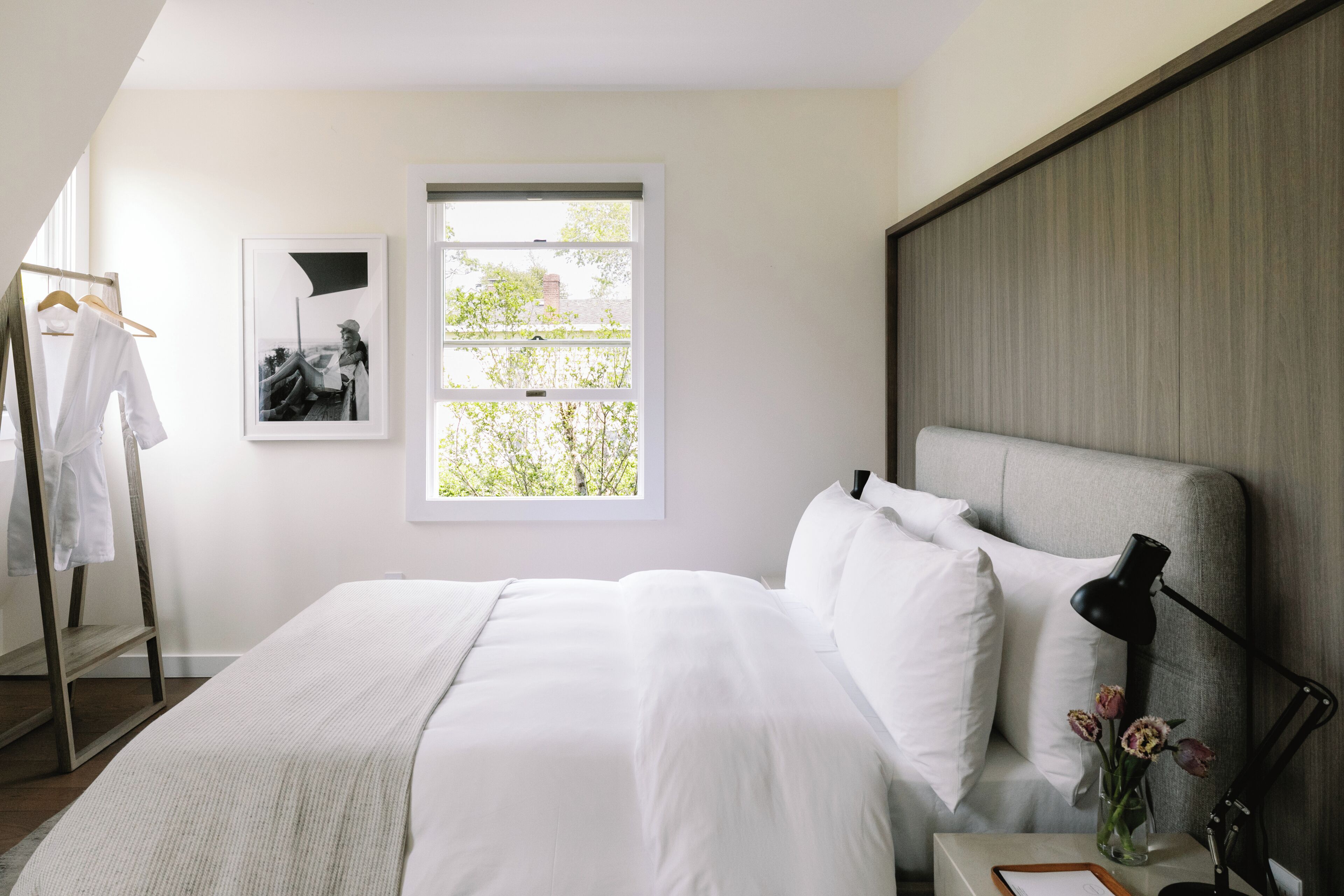 Un dormitorio ordenado con una cama grande, una mesita de noche con lámpara, un cuadro enmarcado en la pared y una ventana con vistas a zonas verdes.