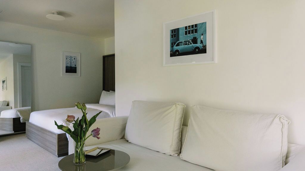 A modern hotel room with a white bed, a white sofa, a small round table with a vase of flowers, and a framed picture on the wall.