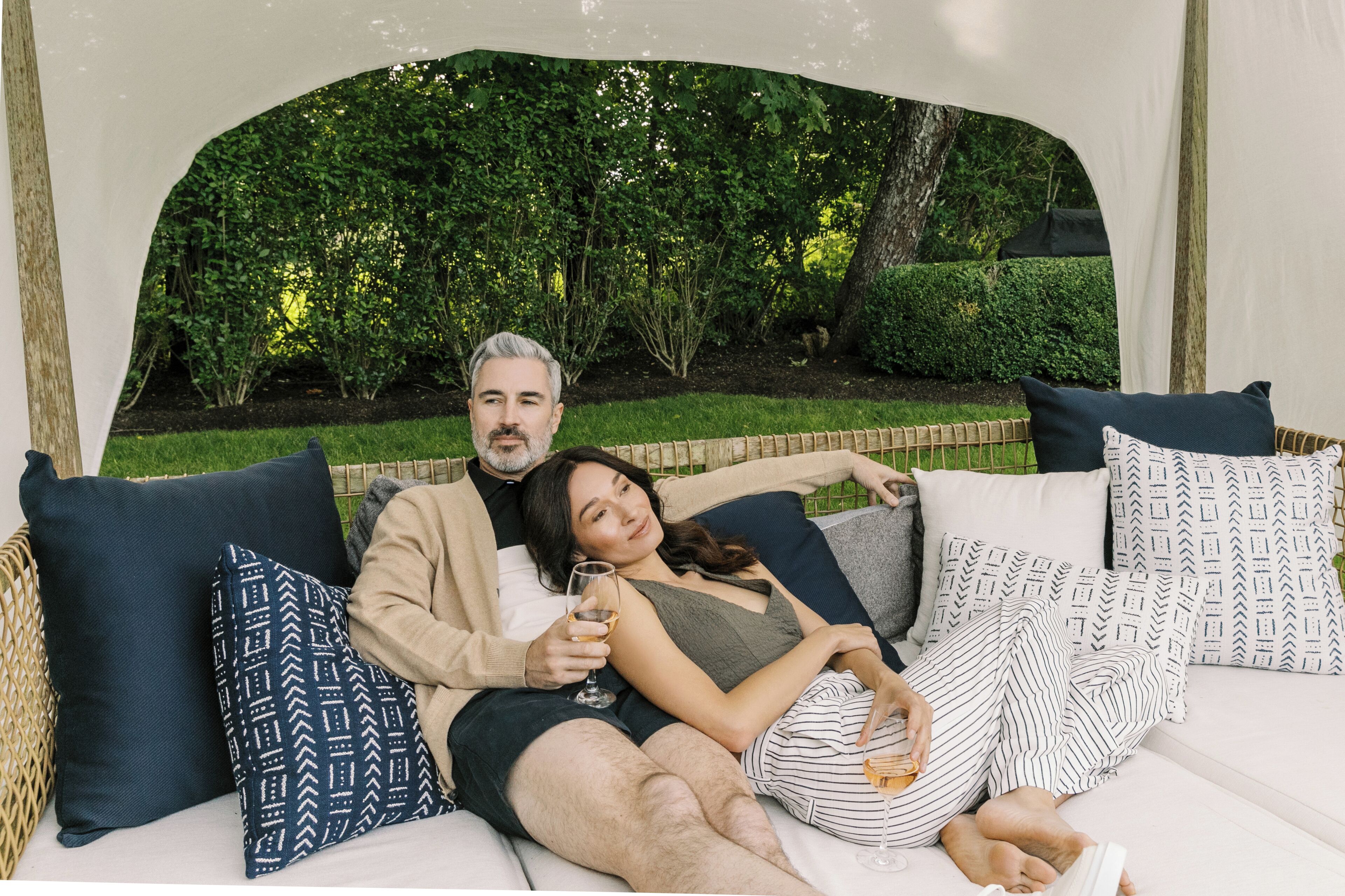 A couple relaxing on a white outdoor sofa with patterned cushions, surrounded by greenery.