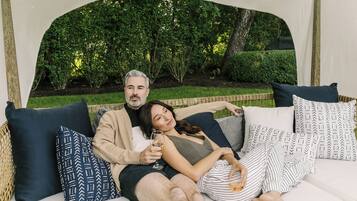 A couple relaxing on a white outdoor sofa with patterned cushions, surrounded by greenery.