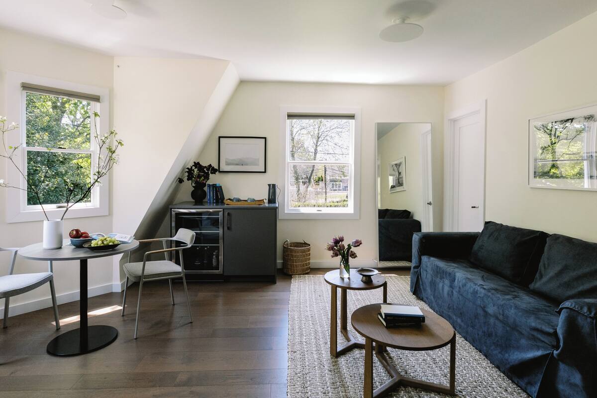 A modern living room with a dark blue sofa, a round coffee table, and a dining area with a white table and chairs. A kitchen with a dark cabinet and a window is visible in the background.