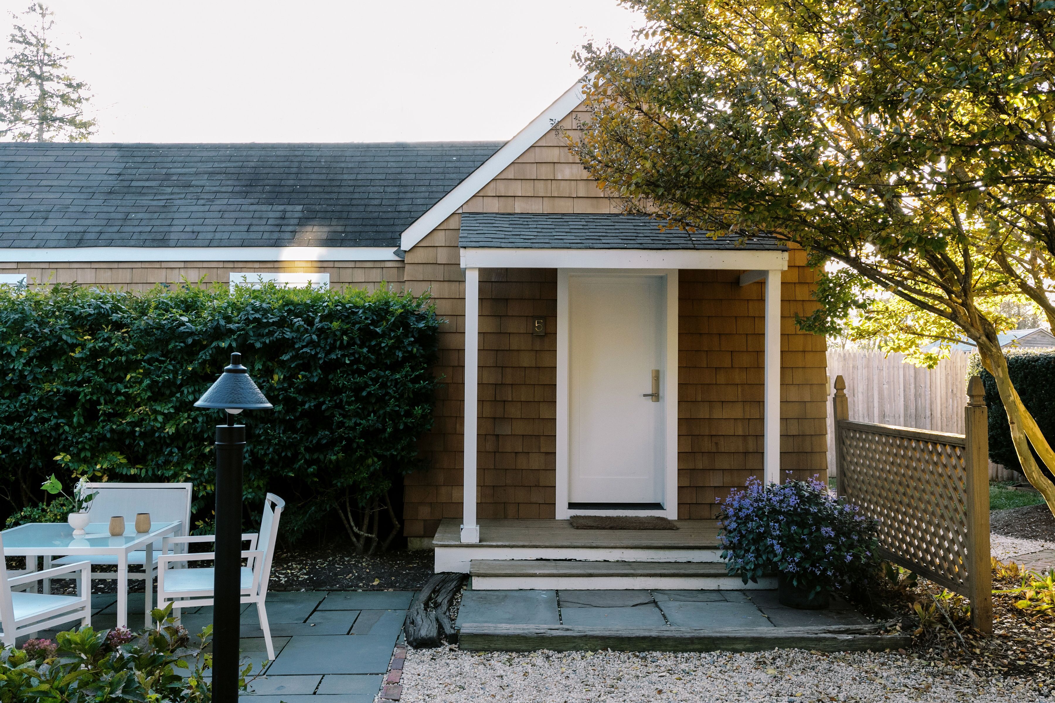 A small house with a wooden exterior, a white door, and a porch with steps. There is a table and chairs outside, and a lamp post is visible.