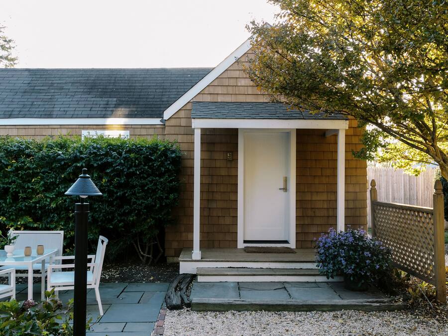A small house with a wooden exterior, a white door, and a porch with steps. There is a table and chairs outside, and a lamp post is visible.