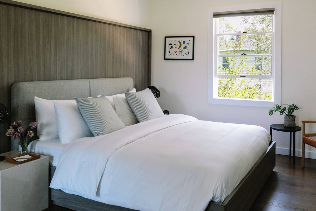A modern bedroom with a large bed, a wooden headboard, a bedside table with a lamp, and a window with a view of trees.