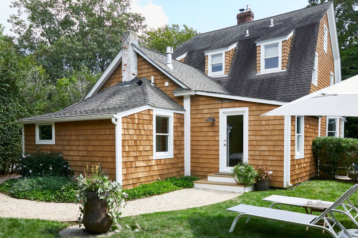 A two-story house with a sloped roof, a front porch, and a small garden area with a bench and potted plants.