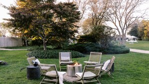 A patio with four wicker chairs arranged around a round table with a fire pit in the center, set in a grassy outdoor area with trees and a fence in the background.