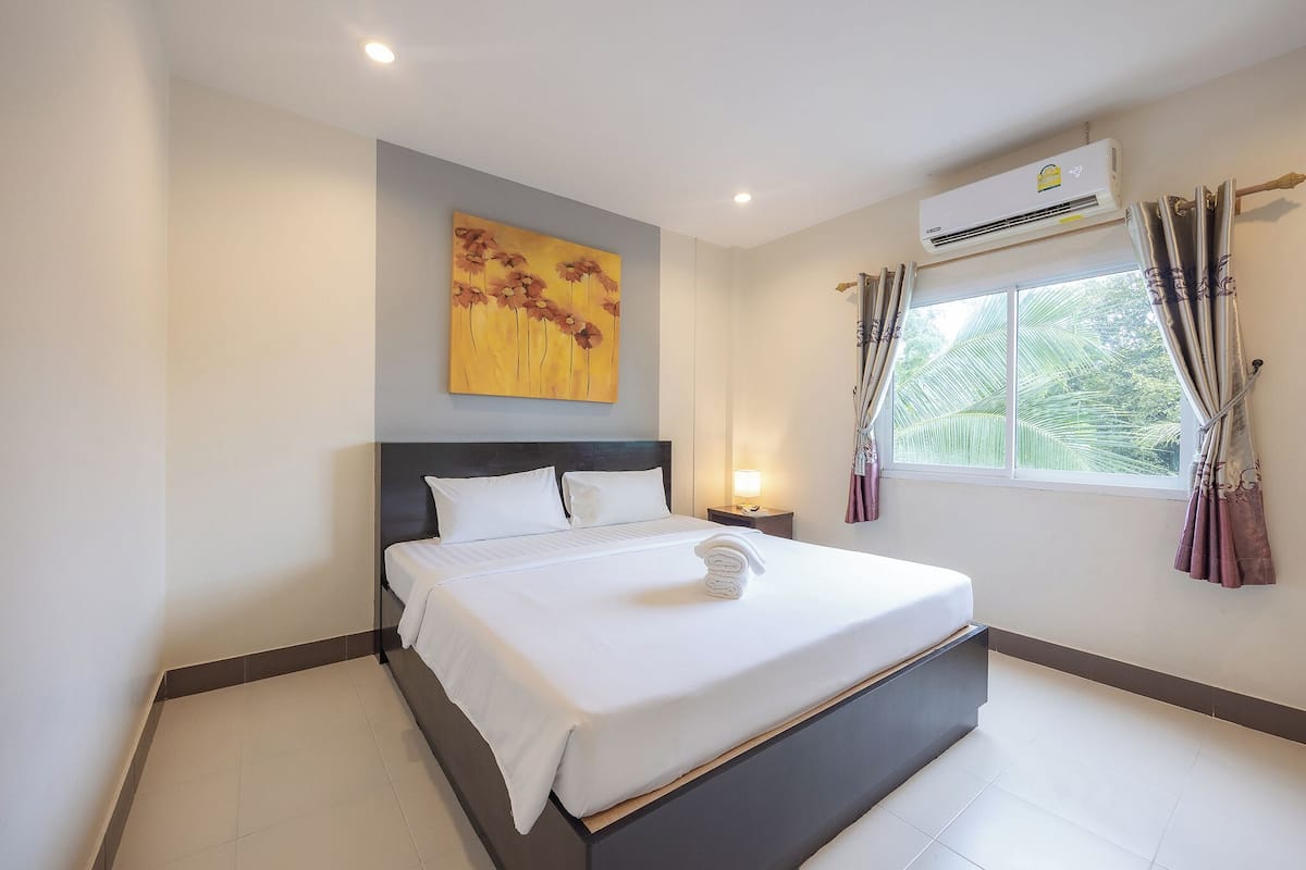 A hotel room with a large bed, a wall-mounted air conditioner, a window with curtains, and a painting on the wall.