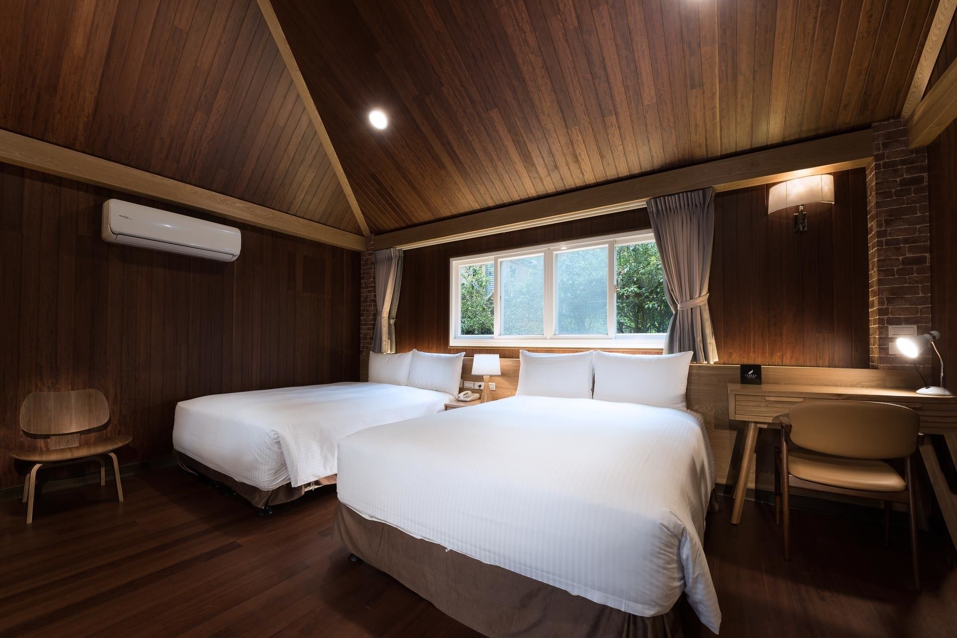 Habitación de hotel con dos camas, un escritorio, una silla y un ventanal con cortinas.