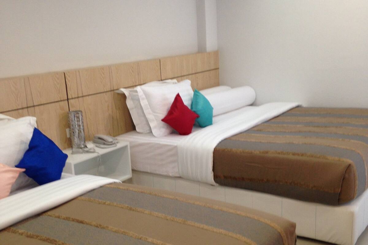 Habitación de hotel con dos camas, cada una con cubrecama a rayas marrones y ropa de cama blanca. Cuenta con un cabecero de madera, una mesita de noche con lámpara y un pequeño atril con teléfono.