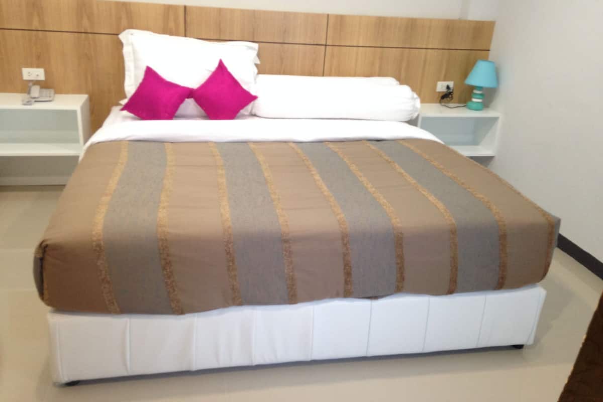 Una cama bien hecha con una colcha a rayas, un almohadón blanco y un almohadón rosa decorativo. Se ve una mesita de noche con una lámpara y una pequeña mesa junto a la cama.