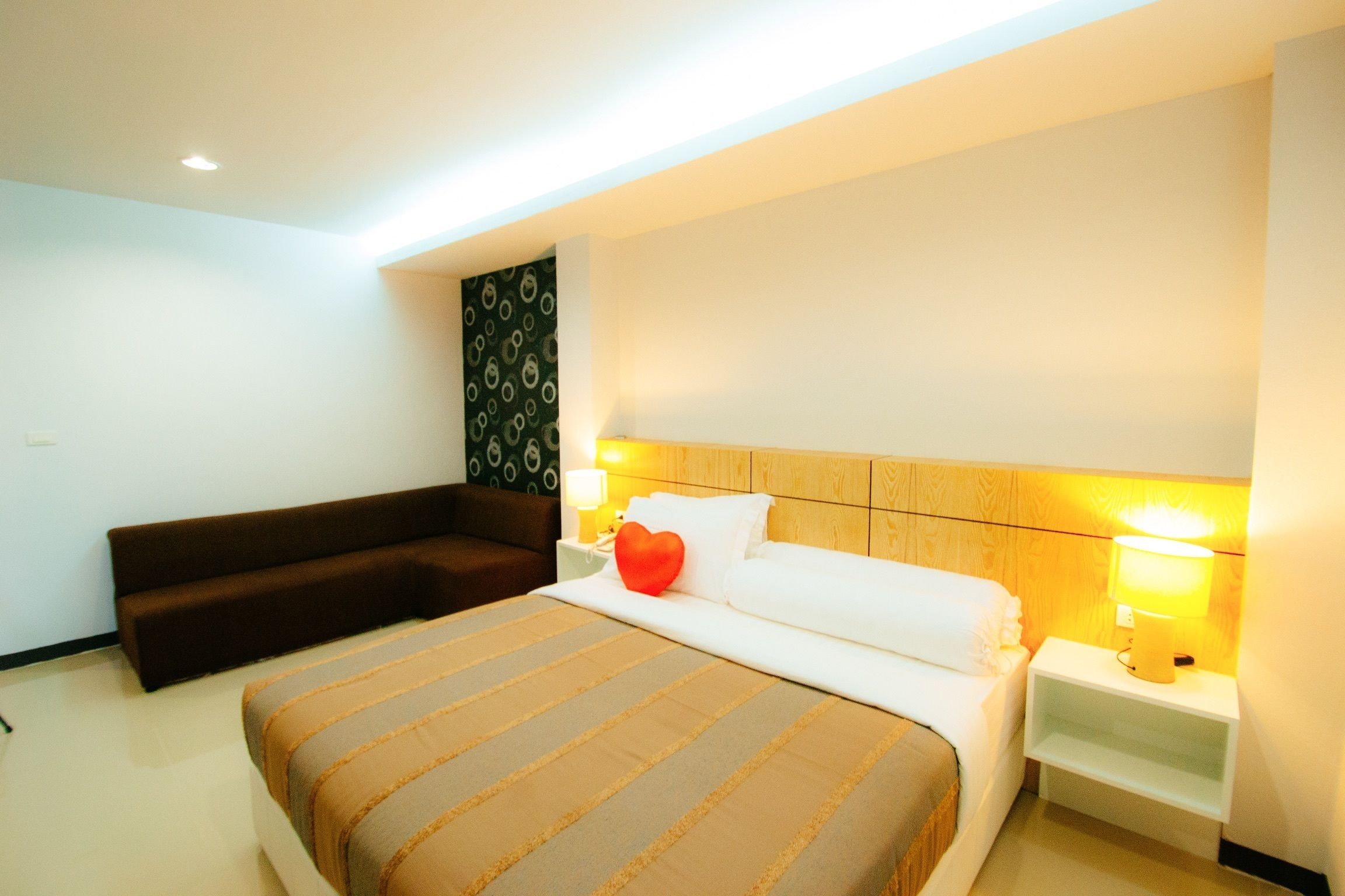 A hotel room with a large bed, a bedside table with a lamp, a sofa, and a patterned wall.