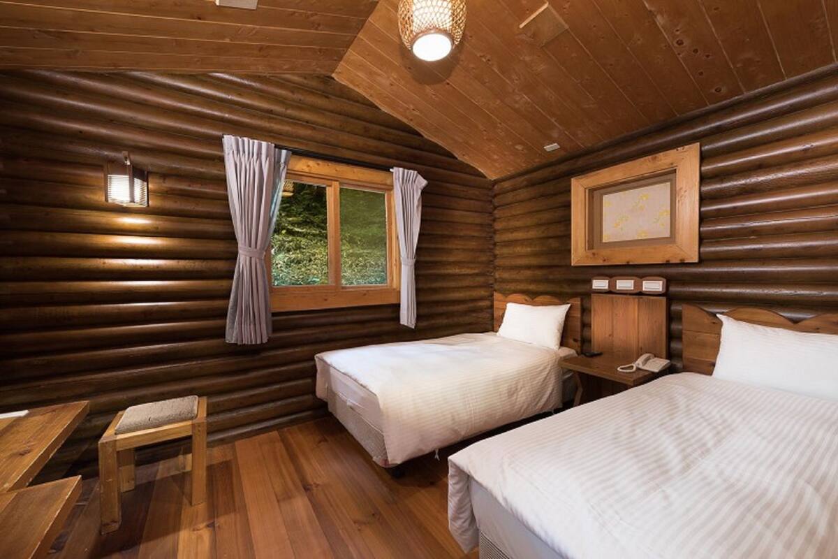 Ein Blockhausräume mit zwei Betten, einem kleinen Tisch und einem Fenster mit Vorhängen.