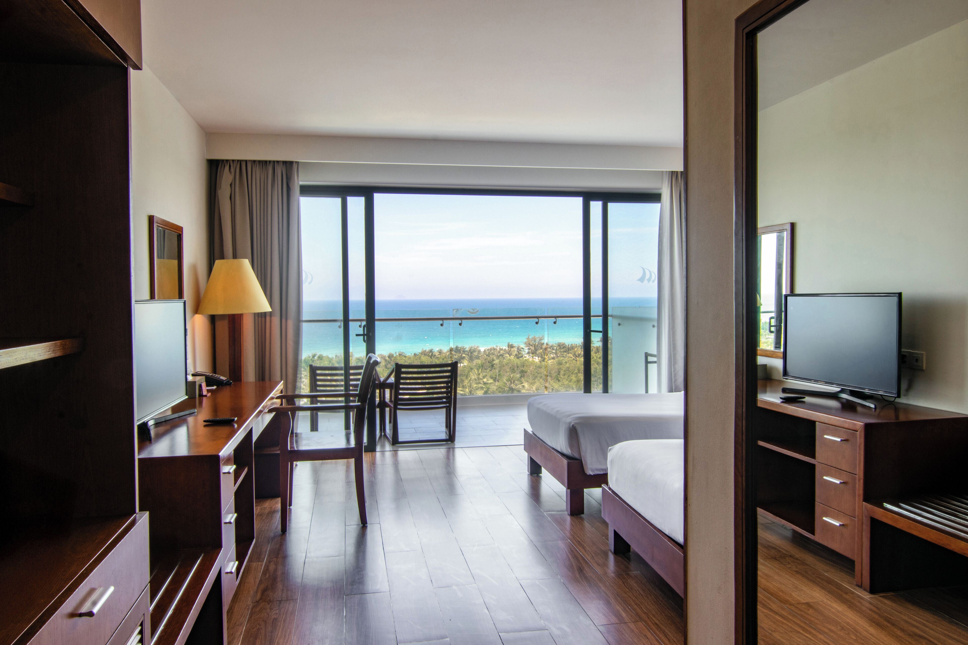 A hotel room with a bed, a desk with a lamp, a TV, and a large window offering a view of the beach.