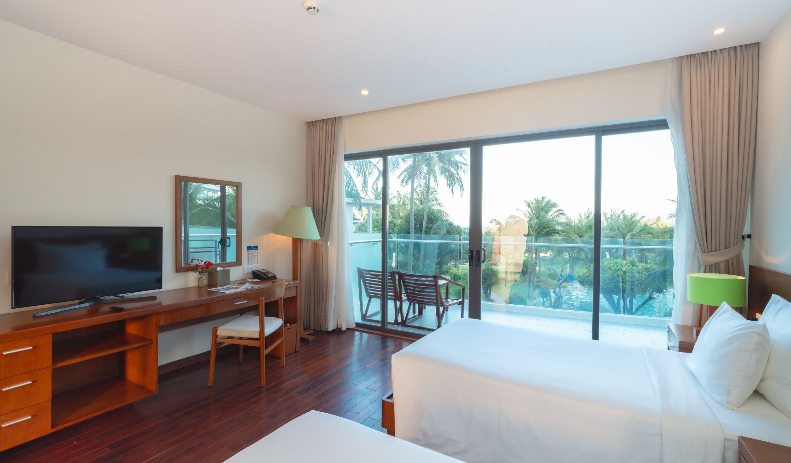 A hotel room with a bed, a desk with a TV, a chair, a balcony with a view of palm trees.