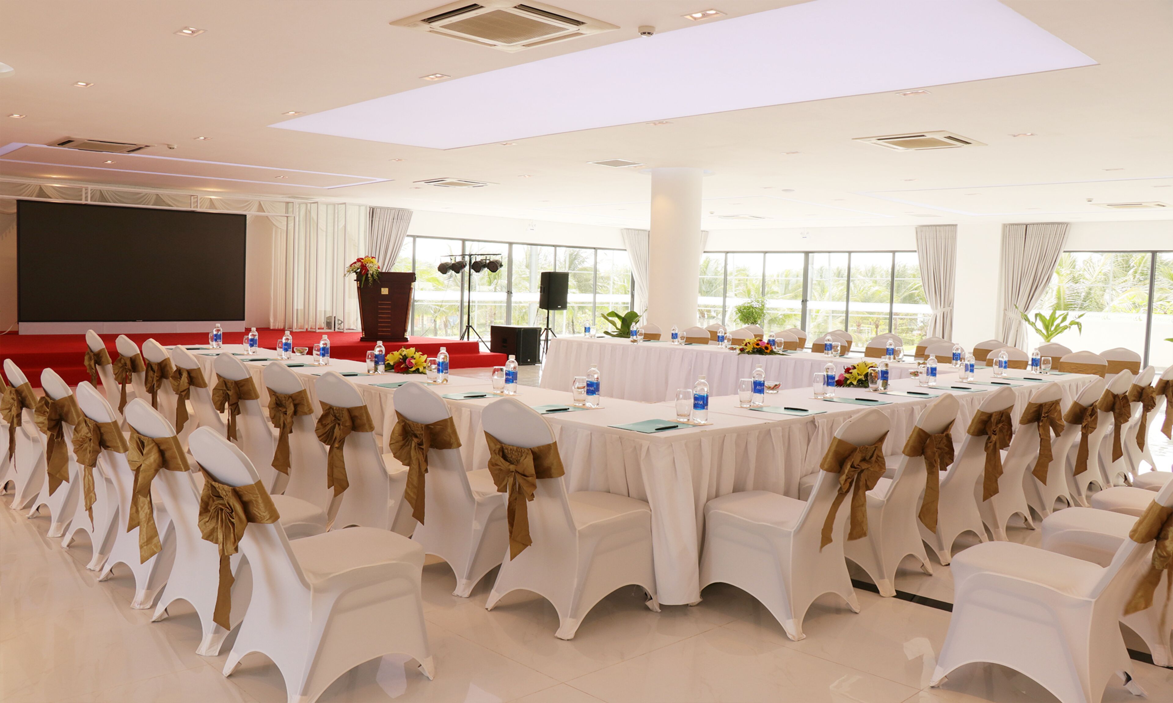 Un salón de conferencias con filas de sillas blancas con cintas doradas, mesas con botellas de agua y un escenario con un atril.