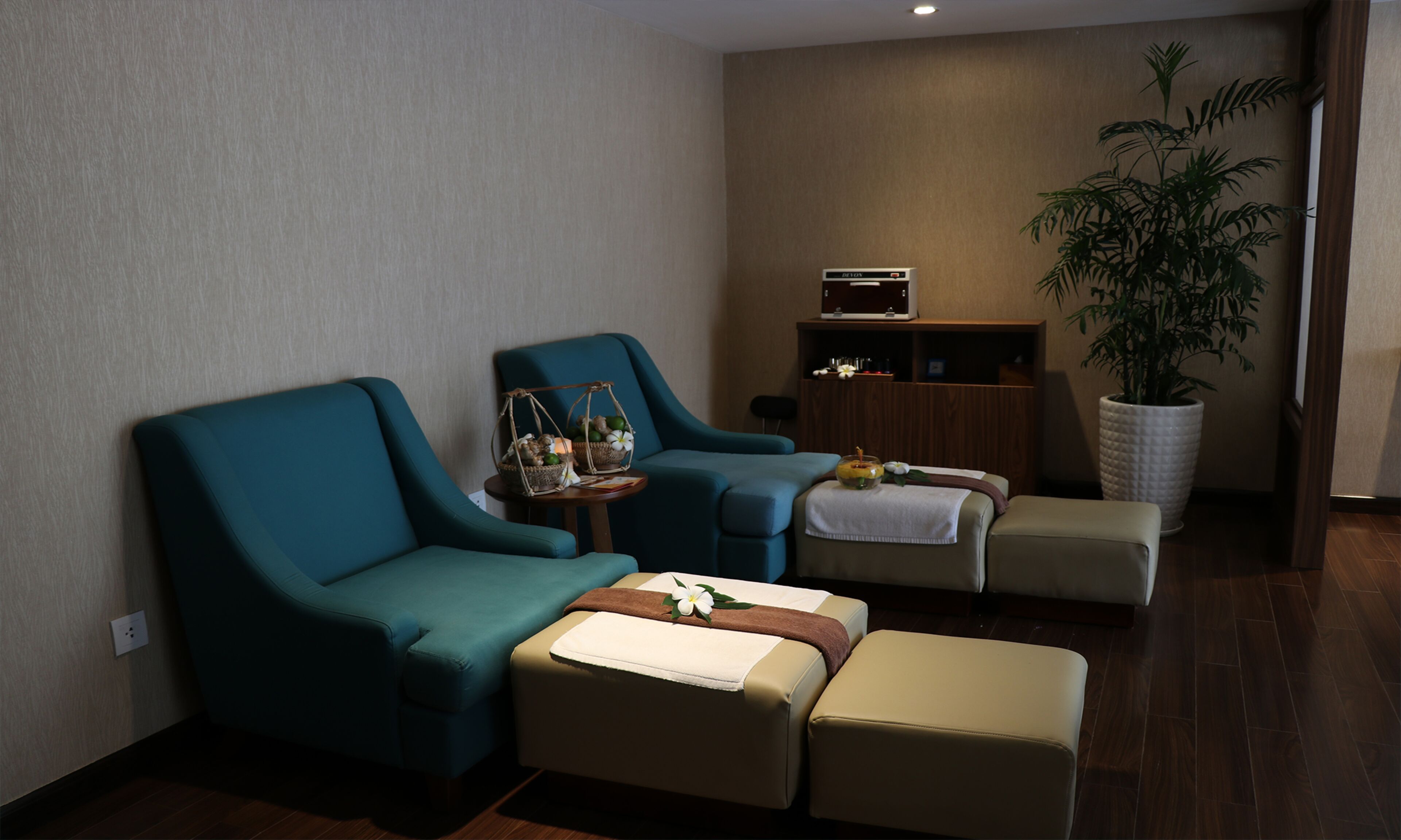 Un cuarto de spa con dos sillones color turquesa, una camilla de masaje, una mesita con una cesta y flores, una planta grande en maceta y un armario de madera.