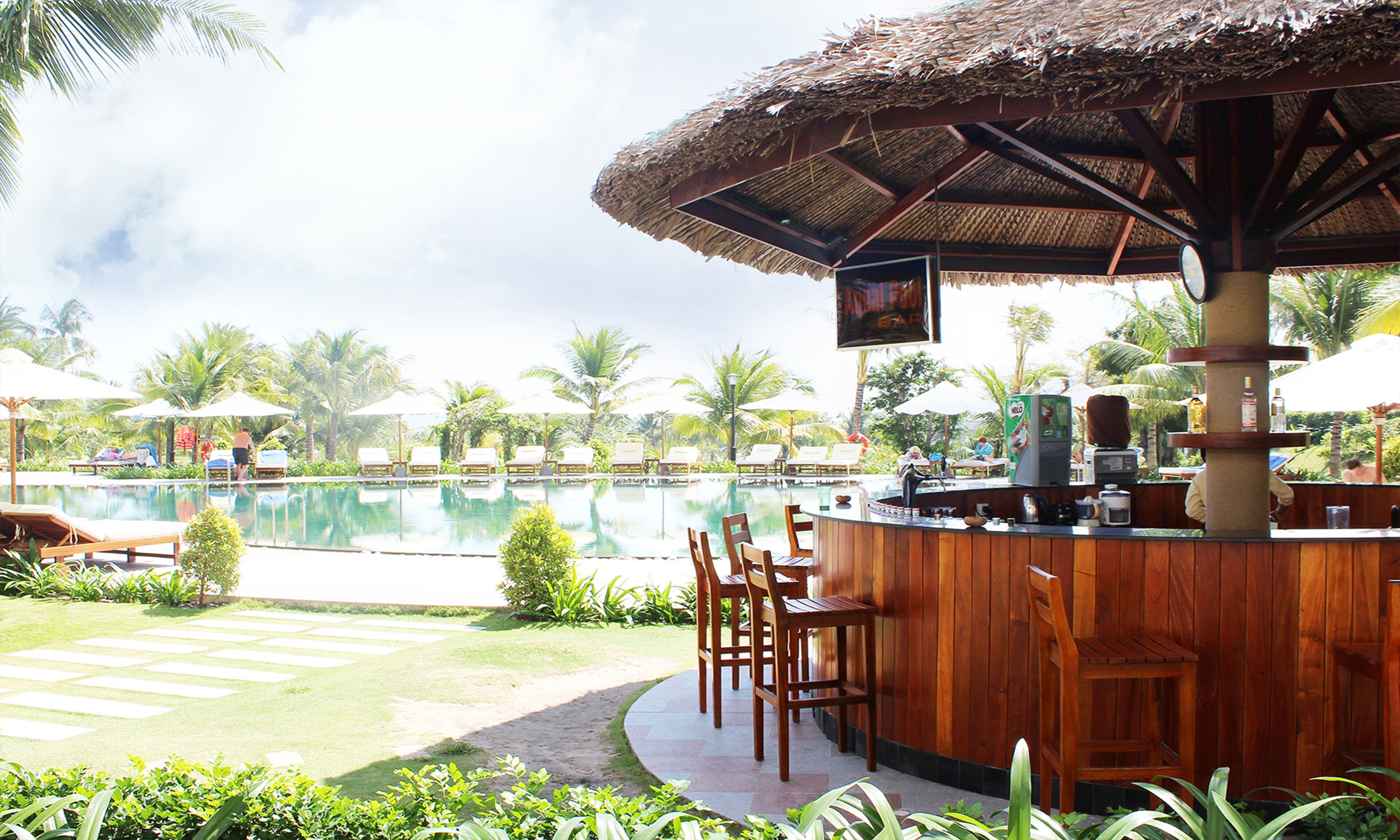 Un bar au bord de la piscine, avec des chaises longues et des parasols, entouré de palmiers et surplombant un ciel dégagé.