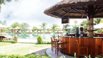 Un bar au bord de la piscine, avec des chaises longues et des parasols, entouré de palmiers et surplombant un ciel dégagé.