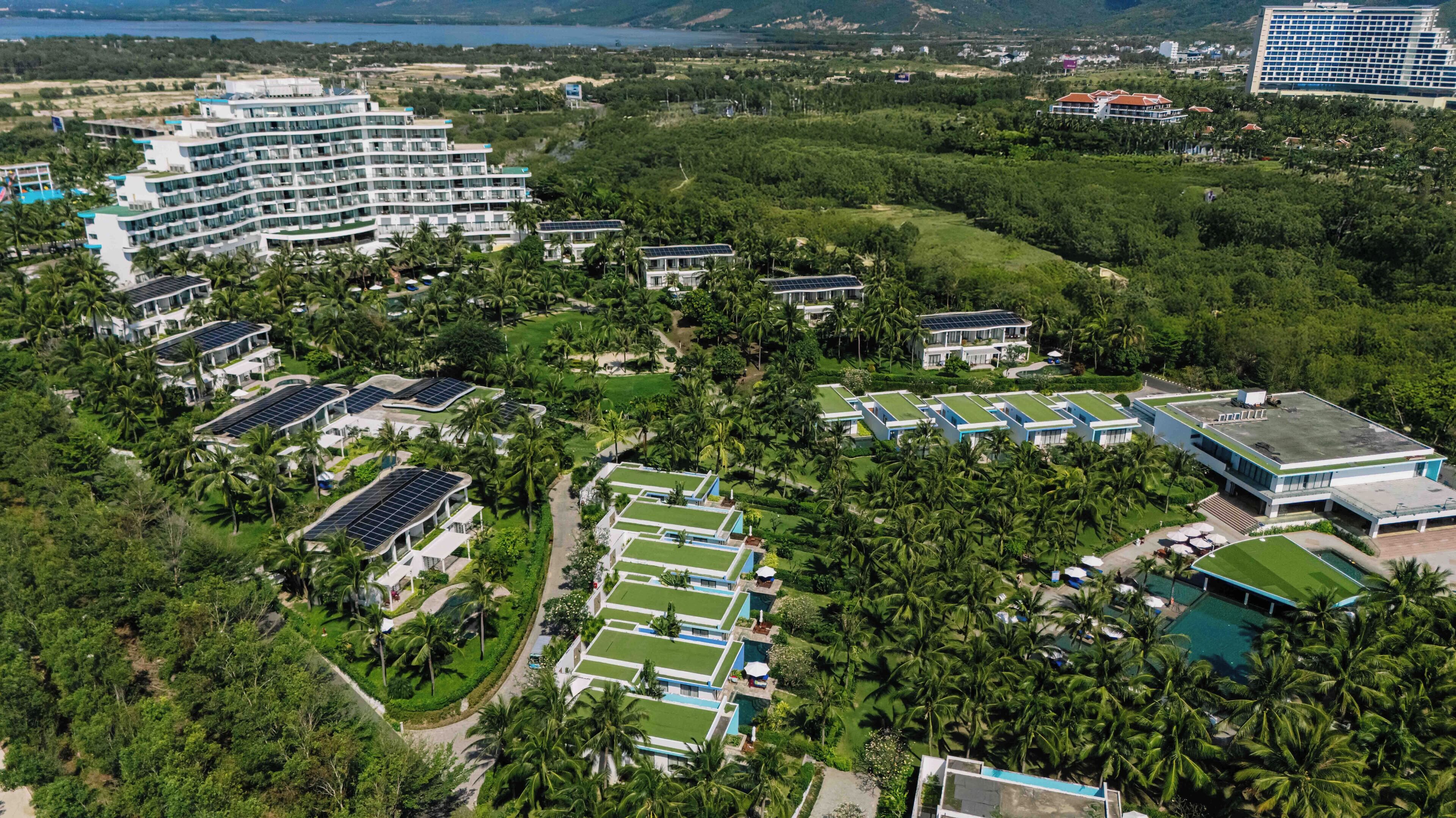 Vue aérienne d’un complexe hôtelier comprenant plusieurs bâtiments, dont un grand hôtel au design moderne, plusieurs villas équipées de panneaux solaires et un espace piscine entouré d’une végétation luxuriante et de palmiers.