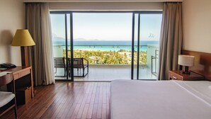 A hotel room with a large bed, a desk, a chair, a lamp, and a balcony with a view of the ocean and distant landscape. - Cam Ranh Riviera Beach Resort & Spa (Cam Lam)