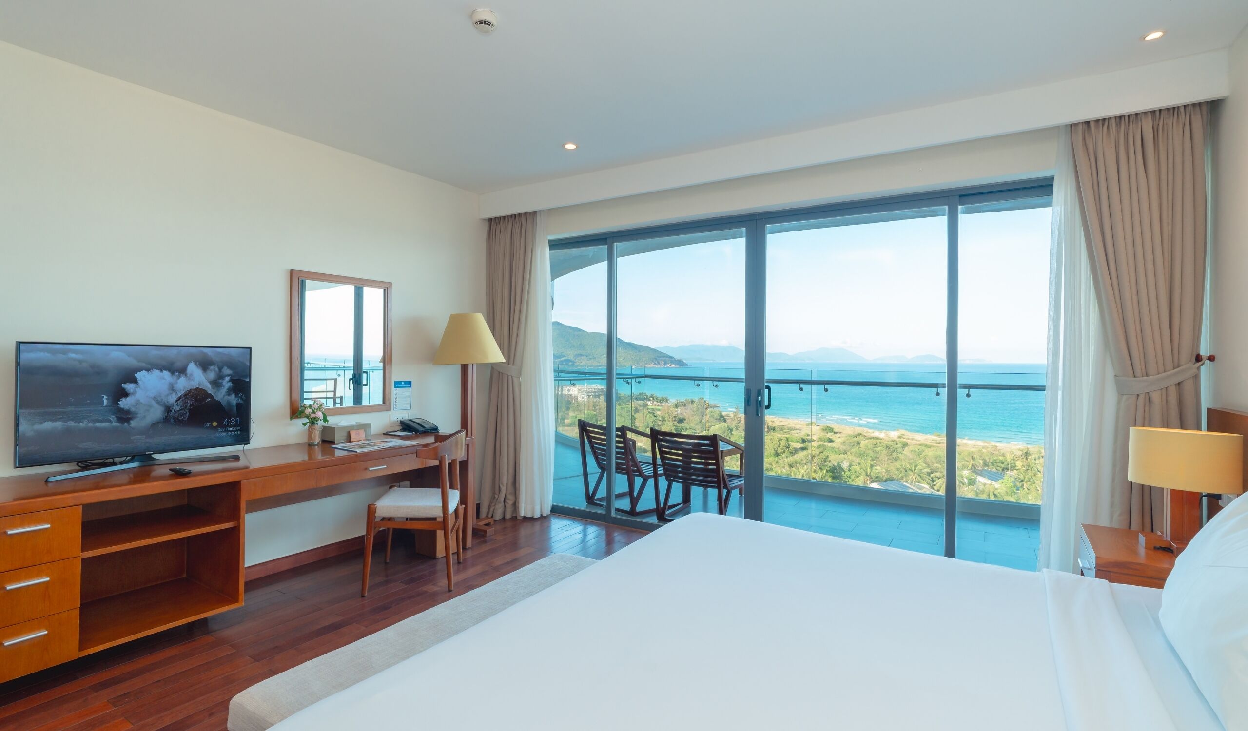 Habitación de hotel con una cama grande, un escritorio con televisión, una silla, un balcón y vistas al mar y las montañas.