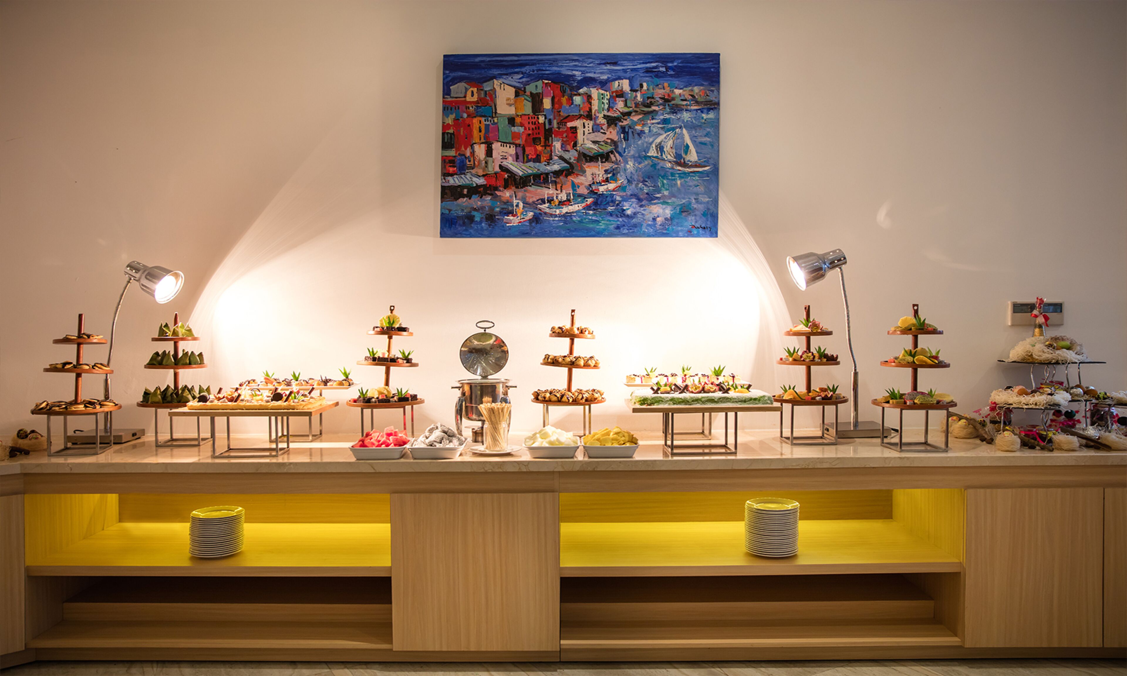 A buffet setup with multiple food stations and a painting of a coastal town on the wall.