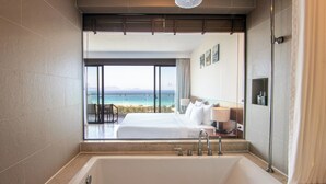 A hotel room with a large bathtub, a bed with white linens, a nightstand with a lamp, and a view of the ocean through large windows. - Cam Ranh Riviera Beach Resort & Spa (Cam Lam)