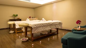 A spa room with two massage tables, a small plant, a bowl of bath salts, and teal chairs.