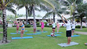 A group of people performing yoga on mats in an outdoor setting with palm trees and a building in the background.