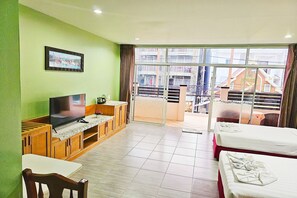 A hotel room with a television, wooden furniture, a dining table, and a balcony with a view of buildings. - Dome Resort (Karon)