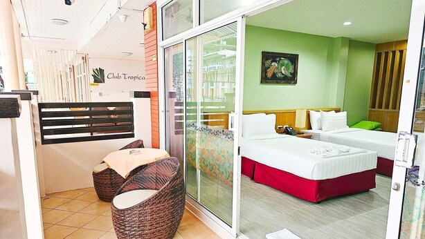A hotel room with a sliding glass door leading to a balcony, a bed with white bedding, a small seating area with wicker chairs, and a green wall with a framed picture. - Dome Resort (Karon)