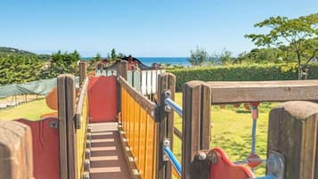 A playground with a slide and climbing area, surrounded by greenery and a view of the sea.