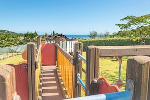 A playground with a slide and climbing area, surrounded by greenery and a view of the sea.