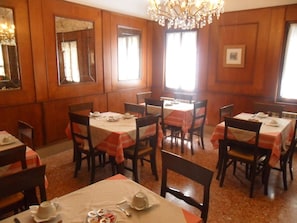 Un comedor con paredes revestidas de madera, ventanas amplias y candelabros. Las mesas están dispuestas con manteles blancos y servilletas anaranjadas.