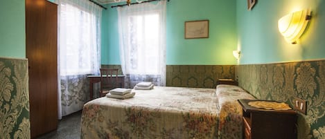 Un dormitorio con cubrecama floral, un candelabro, una mesita de noche de madera y una lámpara fijada a la pared.