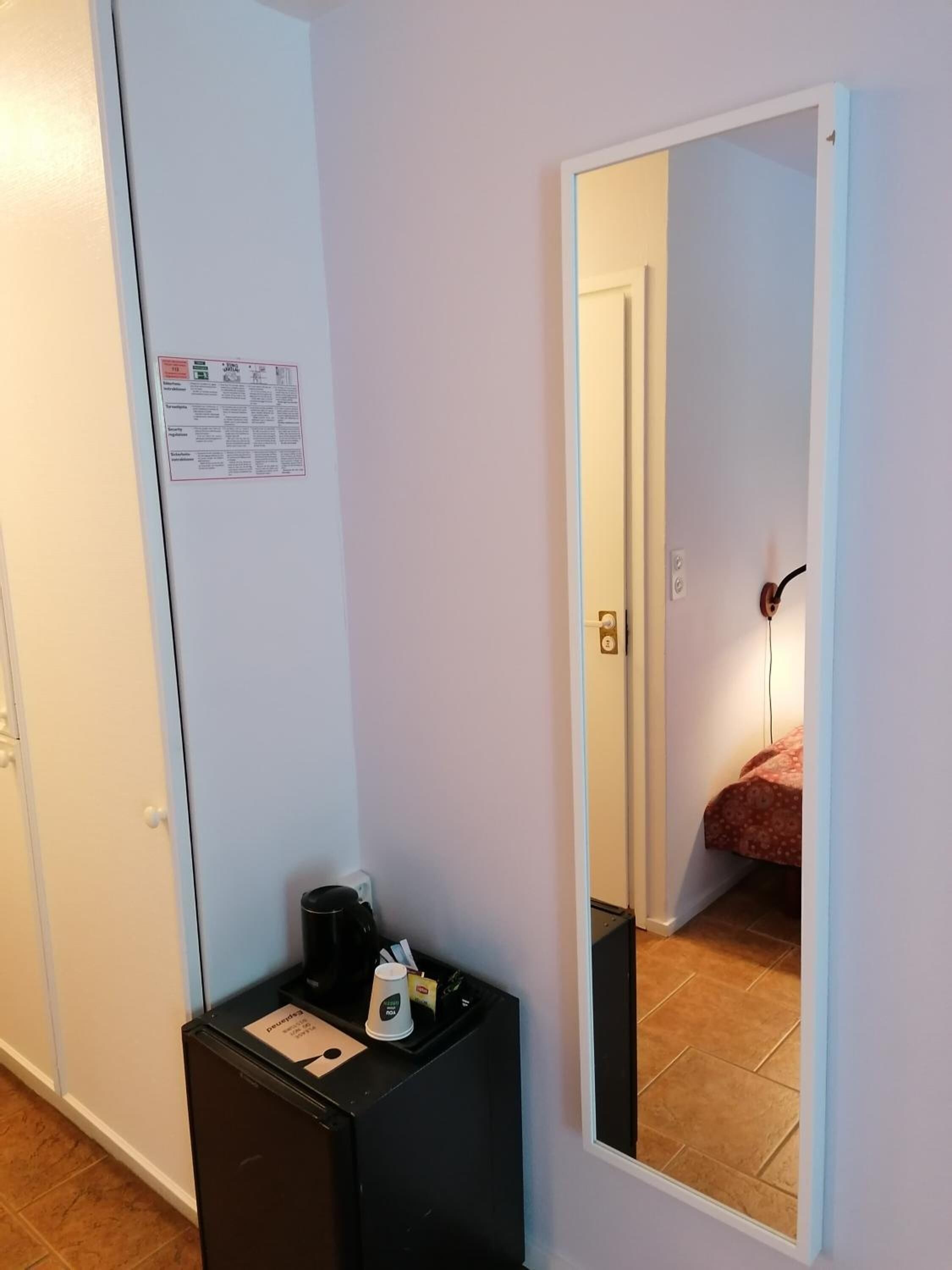 A hotel room with a white door, a small black table with a coffee maker and a cup, and a mirror reflecting a bed with a red blanket.